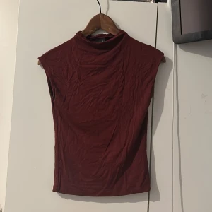 Vinröd topp från Gina Tricot - Vinröd topp från Gina Tricot i softtoutch material. Toppen är ärmlös med hög hals och har en slim passform som sitter snyggt mot kroppen. Perfekt att styla med jeans eller kjol för en clean look. (Finns en liten liten fläck men är ej märkbar när den sitter på) borde gå att få bort med rött tvättredskap