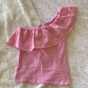 Rosa oneshoulder topp från Kappahl - Supersöt rosa oneshoulder topp från Kappahl med bred volang längs halsringningen. Toppen är ärmlös och har en snygg asymmetrisk design som ger en trendig vibe. Perfekt för dig som gillar färg och vill sticka ut lite extra i sommar. Skit snygg!!! Storlek 158/164, men jag brukar ha xs/s o den passar mig! Hör av er vid frågor!💞😇