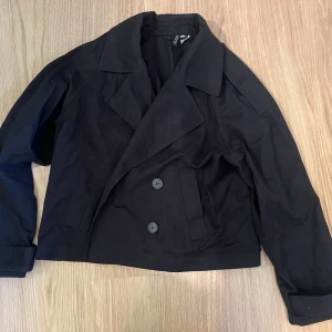 Svart kappa från H&M - Säljer en svart kappa från H&M med klassisk krage och dubbelknäppning framtill. Jackan har två fickor och är tillverkad i bomullsmaterial. Den har en kortare, boxig passform och långa ärmar. Perfekt för dig som gillar stilrena och tidlösa plagg. 