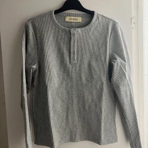 Anovair Henley - Säljer en grå ribbad långärmad tröja från Anovair i storlek S. Tröjan har rund halsringning, knappar framtill och är tillverkad i mjuk bomull med lite stretch. Perfekt för lager-på-lager och chill dagar. Aldrig använd då den är för liten o glömt skicka tbx! Nypris runt 1000kr. 