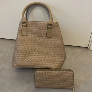 Beige handväska & plånbok Michael Kors - Säljer en beige handväska och matchande plånbok Michael Kors med MK-logga över hela ytan. Väskan har två rejäla handtag, dragkedja och detaljer i ljusbeige skinnimitation. Plånboken har dragkedja i guld och samma mönster.
