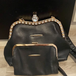 Svart handväska med strass och matchande plånbok - Säljer en svart handväska i skinnimitation med guldfärgade detaljer och strass längs öppningen, samt en stor kristall på låset. Tillhörande svart plånbok med guldfärgad ram ingår. Perfekt för dig som gillar bling och klassisk stil.