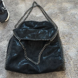 Svart axelväska med kedjedetaljer - Säljer en svart axelväska med coola silverfärgade kedjor som axelrem och längs kanterna. Väskan har en mjuk, lite glansig yta och är sydd med dekorativa sömmar runtom. Perfekt för dig som gillar en edgy och trendig look.