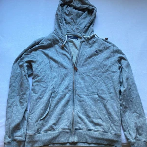 Grå hoodie från Morris med dragkedja - Grå hoodie från Morris i mjuk bomull med hel dragkedja och huva med snörning. Broderad mörkblå logga på bröstet och två stora fickor framtill. Perfekt för chill dagar och enkel att matcha med jeans eller joggers.