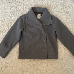 Grå kavajjacka från Gina Tricot - Stilren grå kavajjacka från Gina Tricot med klassisk krage och tre knappar framtill. Jackan har rak passform och är helfärgad utan mönster. Perfekt för dig som gillar en clean och enkel look.