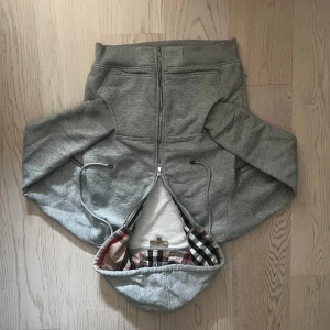 Grå hoodie från Burberry med dragkedja - Snygg grå hoodie från Burberry med hel dragkedja, broderad logga på bröstet och klassiskt rutigt foder i huvan. Mjuk insida och justerbar huva med snörning. Perfekt för dig som gillar stilren streetwear med lyxig touch.