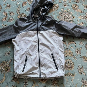 Nike vindjacka med huva, M - Nike vindjacka i grått och vitt med rutigt mönster. Jackan har huva, hel dragkedja framtill och två fickor med dragkedja. Lätt och smidig design, perfekt för blåsiga dagar. Sportig look med svart detalj på fickorna och elastisk nederkant.