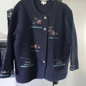 Svart kofta med broderade blommor - Svart kofta från Resis of Sweden i storlek M. Koftan har broderade blommor i blått och grönt på framstycket, fickorna och ärmsluten. Rund hals, dekorativa knappar och två praktiska fickor framtill. Materialet är stickat och känns mjukt och varmt.