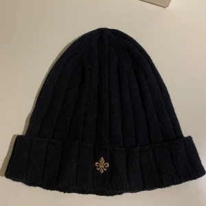 Morris mössa använd 10 ggr - Marinblå ribbstickad mössa med uppvikt kant och en liten guldfärgad broderad lilja framtill. Perfekt för kalla dagar och enkel att matcha med olika outfits.