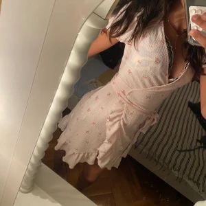 Ljusrosa omlottklänning Gina Tricot XS - Supersöt ljusrosa omlottklänning från Gina Tricot i storlek XS. Klänningen har korta ärmar, volangkant och knyt i midjan. Materialet är mjukt och har små blommiga mönster i rosa och grönt. Perfekt för sommarens alla tillfällen!