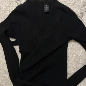 Svart omlott kofta från Soaked in Luxury - Säljer en svart omlott kofta från Soaked in Luxury i storlek S. Tröjan har ribbad struktur, v-ringad hals och långa ärmar. Perfekt för dig som gillar stilrena och enkla plagg med snygg passform. Sitter snyggt och är lätt att matcha med dina favoritjeans.