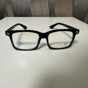 Chrome hearts glasögon - Stilrena svarta glasögon från Chrome Hearts med markerad, fyrkantig båge i blank plast. Utsmyckade med silvriga korsdetaljer i hörnen och på skalmarna samt små stenar längst sidorna. Perfekt för dig som vill sticka ut med en edgy vibe.