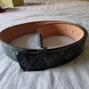 Fransk Bälte 115cm - Snyggt svart Franskt-bälte i skinn med klassiskt monogrammönster och stor logga på spännet. Bältet har en bred design och insidan är ljusbrun. Perfekt accessoar för att lyfta din outfit med exklusiv känsla.