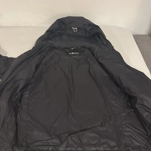 Svart pufferjacka med huva - Svart pufferjacka från Soul River med huva och dragkedja framtill. Jackan har en enkel, stilren design och är vadderad för extra värme. Perfekt för kyliga dagar och har ribbade muddar vid ärmsluten för en skön passform.