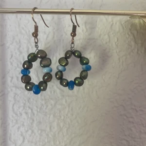 Handgjorda örhängen med äkta pärlor - Unika örhängen i ringform med skimrande mörkgröna pärlor och inslag av blåa och ljusblå pärlor. Perfekta för dig som vill sticka ut med färg och personlighet. Hängande modell med metallkrok.