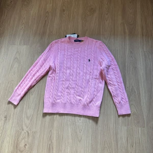 Rosa tröja från Polo Ralph Lauren - Supersöt rosa kabelstickad tröja från Polo Ralph Lauren med klassisk rund halsringning och broderad logga på bröstet. Långärmad modell i mjuk bomull, perfekt för lager-på-lager och chill vibes. Är L men passar som M