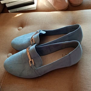 Blå mockaloafers med gulddetalj - Snygga blå loafers i mocka med guldfärgad metallspänne över vristen. Skorna har rund tå och platt sula, perfekt för en stilren och avslappnad look. Passar till både jeans och kostymbyxor.