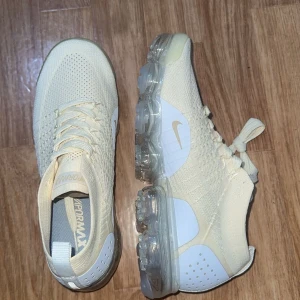 Nike Vapormax ljusa sneakers - Nike Vapormax sneakers i en ljus beige ton med stickad ovandel och transparent sula. Snyggt diskret Swoosh på sidan och klassisk Vapormax-logga på plösen. Skorna har snörning och en modern, sportig vibe som passar till streetwear. Ovandelen är i flyknit-material för extra komfort.