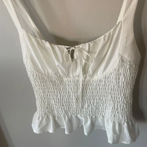 Vit smockad linnetopp från Topshop - Söt vit topp från Topshop med breda axelband, volangkant nedtill. Toppen har knytning framtill och är i storlek 42, M/L. 30kr+frakt
