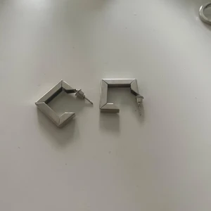 Silvriga kantiga örhängen - Säljer ett par unika silvriga örhängen med kantig, geometrisk form. De är fyrkantiga med en cool, modern design som verkligen sticker ut. Perfekta för dig som gillar statement-smycken och vill ha något annorlunda i smyckeslådan.