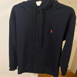 Mörkblå hoodie från Ralph Lauren - Snygg mörkblå hoodie från Ralph Lauren med klassisk röd logga broderad på bröstet. Tröjan har huva med snörning, känguruficka och långa ärmar. Perfekt för en chill och stilren look.