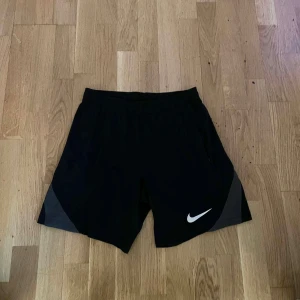 Svarta Nike Dri-FIT träningsshorts - Svarta träningsshorts från Nike med Dri-FIT-teknologi. De har elastisk midja, diskreta grå detaljer på sidorna och en vit Nike-logga framtill. Perfekta för dig som vill ha bekväma och snygga shorts till gymmet eller löprundan.