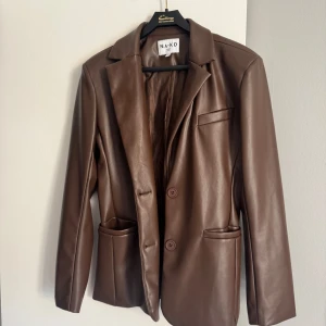 Brun skinnkavaj från NA-KD, strl 38 - Snygg brun kavaj i skinnimitation från NA-KD med klassisk krage, två knappar framtill och tre fickor. Stilren och rak modell med slät yta och fodrad insida. Perfekt för dig som gillar en cool och trendig look.