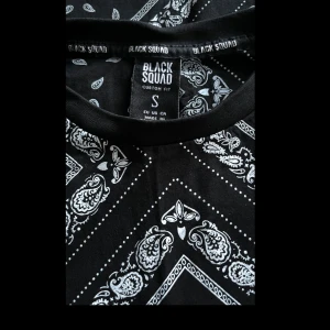 Svart bandana-mönstrad t-shirt Black Squad - Svart t-shirt från Black Squad i storlek S med grymt bandana-inspirerat vitt mönster över hela tröjan. Klassisk rund halsringning och korta ärmar. Skön passform och riktigt snygg streetstyle-vibe. Perfekt för dig som vill sticka ut lite extra.