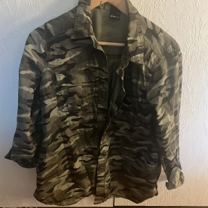 Camouflage overshirt från Gina Tricot - Säljer en snygg overshirt från Gina Tricot i camouflage-mönster. Skjortan har klassisk krage, knäppning framtill och två bröstfickor. Perfekt att slänga över en t-shirt för en cool look. Färgerna går i grönt, beige och svart.