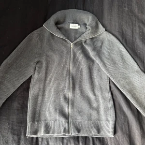 Grå stickad kofta från East West - Säljer en grå stickad kofta från East West i storlek M. Koftan har hel dragkedja framtill, ribbad struktur och hög krage. Perfekt för lager-på-lager och funkar till både jeans och chinos. Enkel och stilren design som passar till många outfits.