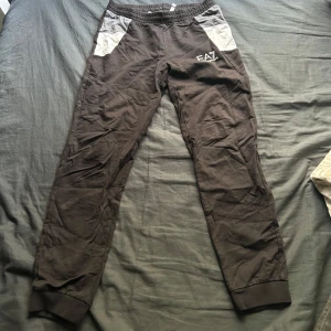 Svarta EA7 mjukisbyxor med grå detaljer - Snygga svarta mjukisbyxor från EA7 Emporio Armani med grå paneler vid fickorna och logga på låret. Byxorna har resår i midjan och muddar vid bensluten för en skön passform. Perfekta för chill eller träning.