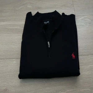 Svart half zip tröja från Polo Ralph Lauren - Svart långärmad tröja från Polo Ralph Lauren med klassisk röd broderad logga på bröstet. Tröjan har en snygg half zip-dragkedja och hög krage. Perfekt för dig som gillar stilren och tidlös design.