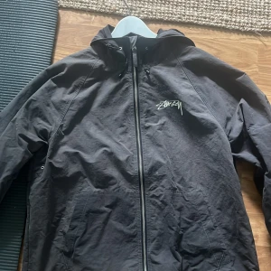 Mörkgrå vindjacka från Stüssy, M - Svart vindjacka från Stüssy med ikonisk logga på bröstet och stor logga på ryggen. Jackan har huva med snörning, dragkedja framtill och två fickor. Perfekt för streetwear-stil och enkel att slänga på sig när det blåser.