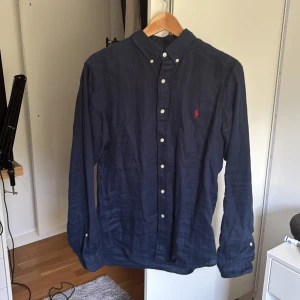 Mörkblå linneskjorta Ralph Lauren M - Snygg mörkblå skjorta från Ralph Lauren i slim fit-modell. Tillverkad i 100% linne med klassisk button-down krage, vita knappar och den ikoniska röda broderade loggan på bröstet. Perfekt för dig som gillar stilrena och tidlösa plagg. Finns qr för att se att den är äkta! Nästan aldrig använd