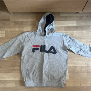 Grå hoodie från FILA med logga - Snygg grå hoodie från FILA med stor logga framtill i svart och rött. Tröjan har huva med mörkblå insida och en klassisk känguruficka. Perfekt för dig som gillar sportig och avslappnad stil.