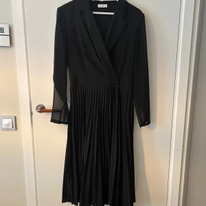 Svart plisserad långklänning Sandro - Elegant svart långklänning från Sandro Paris med plisserad kjol, omlottliv och långa ärmar i lätt, flowigt material. Klänningen har en snygg v-ringning och knäppning i midjan som ger en stilren och chic look.