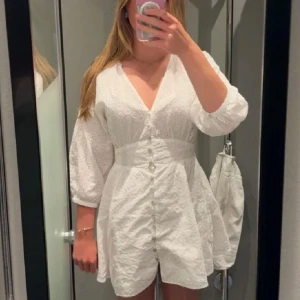 Vit kort klänning med v-ringning - Superfin vit kort klänning med v-ringning och knappar framtill. Klänningen har trekvartsärmar, markerad midja och broderade detaljer i tyget. Perfekt för sommarens alla tillfällen och ger en fräsch och luftig känsla.