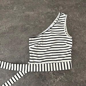 Randig one shoulder topp från NA-KD - Skitsnygg vit och svart randig topp från NA-KD med one shoulder-design, som tyvärr måste säljas då den blivit förliten. Köparen står för frakten!