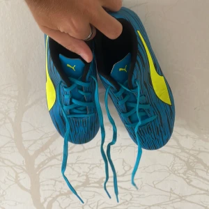 Blå fotbollsskor från Puma - Snygga blå fotbollsskor från Puma med coolt svart mönster och stora gula loggor på sidorna. Skorna har snörning och dobbar för gräsplan. Perfekta för dig som vill sticka ut på planen med stilren design och skön passform. Väldigt bekväma på planen och skorna är knappt använda. ABS! Pris går att diskutera.