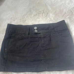 Svart jeanskjol med fickor - Svart kort jeanskjol med två framfickor, två bakfickor och dubbla silverknappar i midjan. Kjolen har bälteshällor och klassisk jeansdesign, perfekt för en cool och enkel stil. Materialet är stadigt och har en snygg passform. Osäker på storlek men xs-s