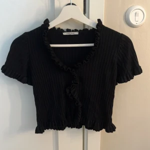 Svart ribbad blus från Chiquelle - Supersöt svart ribbad blus från Chiquelle med volangkant längs halsringning, knäppning framtill och korta puffärmar. Croppad modell med stretchigt material och fina detaljer som ger en trendig look. Perfekt att styla med jeans eller kjol.storlek är jag inte säker på men skulle tippa på xs/s❤️❤️