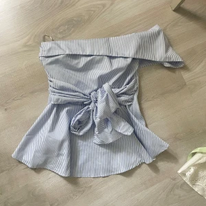 Randig offshoulder topp med knyt - Superfin blåvit randig offshoulder topp med bred volang upptill och stor knytning i midjan. Toppen har en figurnära passform upptill och faller ut nertill för en snygg siluett. Perfekt till jeans eller kjol för en trendig look.