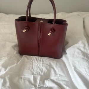 Vinröd handväska med gulddetaljer - Snygg vinröd handväska i skinn med två handtag och guldfärgade detaljer. Väskan har dragkedja upptill och en knappstängning. Stilren och kompakt modell som passar perfekt till dina viktigaste grejer.❤️