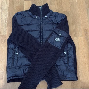 Svart Moncler pufferjacka med stickade ärmar - Snygg svart cardigan från Moncler med stickade ärmar och detaljer. Jackan har dragkedja framtill, hög krage och Moncler-logga på ärmen. Perfekt för dig som vill ha en jacka som sticker ut och känns både sportig och stilren nu till hösten.