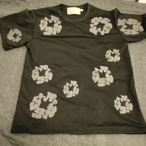 Svart t-shirt Denim Tears med blommor - Svart t-shirt från Denim Tears med gråa blommiga cirkelmönster tryckta över hela plagget. Klassisk passform med rund hals och korta ärmar. Materialet känns mjukt och skönt, perfekt för en avslappnad streetwear-look.