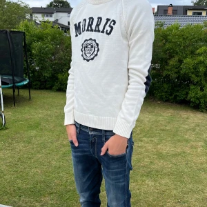 Vit stickad tröja från Morris - Vit stickad tröja från Morris med broderad logga och text på bröstet. Tröjan har svarta armbågslappar, ribbade muddar och rund hals. Perfekt för dig som gillar klassisk och sportig stil.