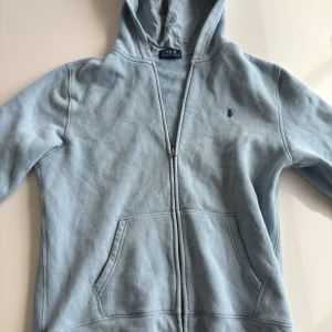 Ljusblå hoodie från Polo Ralph Lauren - Snygg ljusblå hoodie från Polo Ralph Lauren med dragkedja och huva. Broderad logga på bröstet och stort tryck med Polo Ralph Lauren på ryggen. Mjuk bomullskänsla och känguruficka framtill. Perfekt för chill dagar.