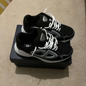 Svarta Dior sneakers med vita detaljer - Snygga svarta sneakers från Dior med vita och grå detaljer samt vita snören. Skorna har en sportig design med rund tå och platt sula. Perfekta för dig som vill ha en exklusiv och trendig look med tydlig logga på sidan.