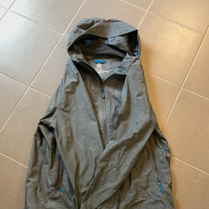 Grå vindjacka från Champion - Snygg grå vindjacka från Champion med huva och vattentät, andningsbar teknologi. Jackan har hel dragkedja framtill, två fickor med dragkedja och blå detaljer. Perfekt för blåsiga dagar och outdoor-äventyr.