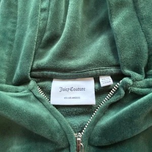Grön velour hoodie från Juicy Couture - Säljer en grön hoodie från Juicy Couture i mjuk velour med dragkedja framtill och broderad JC-logga på bröstet. Tröjan har huva, långa ärmar och två fickor fram. Perfekt för dig som gillar chill och retro vibes.